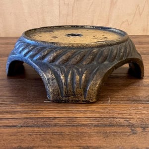 Può includere: Un piedistallo decorativo color bronzo con una parte superiore rotonda e un motivo a foglie sui lati. Il piedistallo ha tre gambe curve e un piccolo foro al centro della parte superiore. Il piedistallo è in metallo.