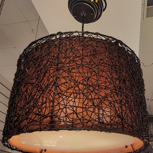 May include: A pendant light fixture with a woven, dark brown shade. The shade has a rounded, bell-like shape and is suspended from a dark metal ceiling mount. The interior of the shade has a warm, orange glow, and the bottom is a white, circular diffuser.
