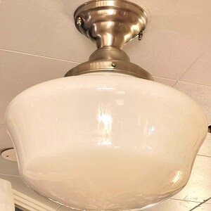 Può includere: Un apparecchio di illuminazione a soffitto in stile vintage con un paralume in vetro bianco e finitura in nichel spazzolato. Il paralume ha una forma arrotondata a forma di ciotola. L'apparecchio è montato a soffitto.