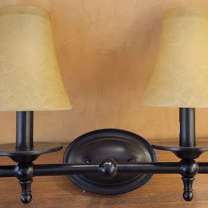 May include: A two-light wall sconce with a dark bronze finish. It features two beige, bell-shaped shades with a textured pattern. The sconce has a horizontal bar with decorative finials and a central oval backplate.