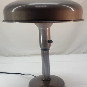 Puede incluir: Lámpara de escritorio de estilo vintage con acabado bronce metálico. La lámpara presenta una pantalla redondeada, un vástago cilíndrico con ranuras verticales y una base circular. Tiene un pequeño botón y un cable de alimentación negro.