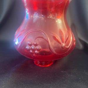 May include: A translucent ruby red glass lamp shade featuring a flared top rim and a rounded body. The exterior surface is decorated with embossed patterns including clusters of grapes and stylized floral motifs, with a circular opening at the base.