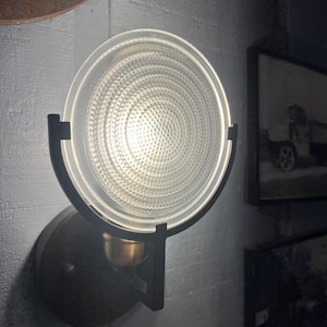 Könnte beinhalten: Eine Wandleuchte mit einem kreisförmigen, strukturierten Glasschirm. Der Schirm wird von einem schwarzen Metallrahmen gehalten und auf einer dunkelbronzenen Basis montiert. Das Licht ist eingeschaltet und wirft einen warmen Schein. Die Leuchte ist an einer grauen Wand befestigt.