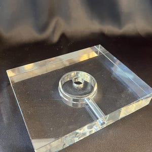 May include: A clear, rectangular acrylic display base featuring a centered, circular raised ring with a small cylindrical protrusion extending from one side, designed to hold or showcase a specific object in a stable, upright position.