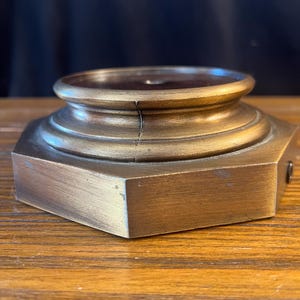 May include: A metallic, bronze-toned display base with an octagonal bottom section and a circular, tiered top platform. The base features a visible vertical crack running through the center of the raised circular portion and a small screw on the side.
