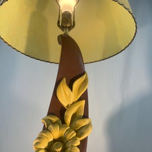Może przedstawiać: Wintage lampa stołowa z żółtym abażurem i brązową podstawą. Podstawa posiada żółty kwiatowy wzór. Lampa ma mosiężny uchwyt na żarówkę i żarówkę. Abażur ma ozdobne wykończenie.