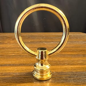 May include: A gold-colored metal finial featuring a circular ring attached to a tiered, cylindrical base. The surface of the object has a polished, reflective finish, and the design consists of a smooth, continuous loop connected to a solid pedestal.