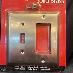 May include: A polished nickel solid brass light switch cover with a toggle switch and a rectangular opening. The cover is packaged on a red and black card with the brand name "Amerelle" and the words "Solid Brass" printed on it.