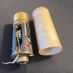 May include: A vintage light bulb socket mechanism with a gold-toned threaded base, a dark gray internal frame, and a silver ball chain pull switch. Beside the socket lies a cylindrical, light-colored cardboard sleeve cover for the fixture.