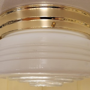 May include: A vintage ceiling light fixture with a gold-colored metal band and a white, ribbed glass shade. The light has a classic design, suitable for various interior styles. The fixture is mounted on a white ceiling.