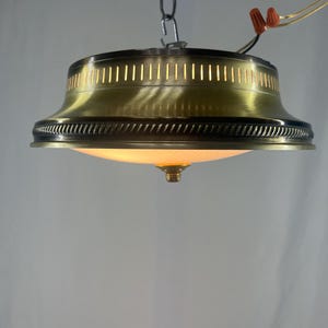 Może przedstawiać: Zabytkowa mosiężna lampa sufitowa z ozdobną krawędzią i kloszem z matowego szkła. Światło zwisa z łańcucha i ma pionowe szczeliny u góry. Oprawa jest podświetlona, ​​emitując ciepłą poświatę.