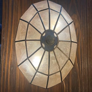 Könnte beinhalten: Eine ovale Buntglaslampe mit geometrischem Design. Die Lampe hat helles, durchscheinendes Glas mit dunklen Linien, die ein Muster bilden. Die Lampe ist auf einer dunklen Holzoberfläche montiert.