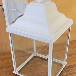 May include: A white outdoor lantern with a classic design. The lantern features a square glass enclosure with a white metal frame and a decorative top. It is designed to be mounted on a wall.