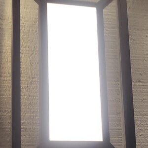 May include: Rectangular black metal light fixture with a bright white illuminated panel. The fixture is mounted on a textured, light-colored wall, creating a modern, minimalist design.