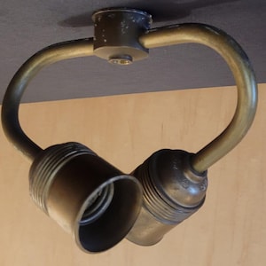 May include: A vintage brass-toned light fixture with two bulb sockets. The fixture has a curved design, with the sockets angled downward. The fixture is mounted on a dark gray surface, with a light wood-toned surface in the background.