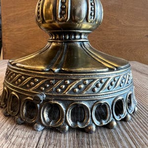 May include: Close-up of an antique brass lamp base. The base features ornate detailing, including a decorative band with small circles and a series of openwork arches. The top portion has a bulbous shape with vertical ridges. The finish has a vintage, aged appearance.