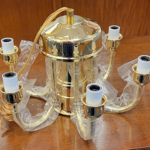 May include: A gold-colored chandelier with five light sockets. The fixture has a central cylindrical body and curved arms extending to hold the sockets. The chandelier is wrapped in plastic and sits on a wooden surface.
