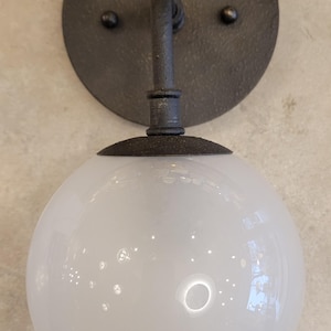 May include: A wall-mounted light fixture featuring a round, dark metal backplate with two small rivets. A curved arm supports a white, spherical glass globe. The fixture has a vintage or industrial aesthetic.