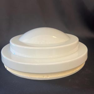 May include: A circular, white ceramic lid featuring a tiered, stepped design with a smooth, rounded dome at the center. The underside of the lid has a thick, unglazed, off-white rim that is designed to fit securely onto a corresponding container.