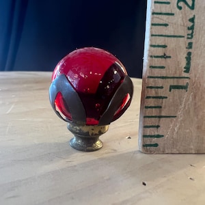 Könnte beinhalten: Ein roter Glaskugel-Endknopf mit einem metallischen Käfigsockel, platziert neben einem Holzlineal, das eine Höhe von etwa 3,8 Zentimetern anzeigt. Das rote Glas weist interne Blasen auf und wird von einem dunklen, geschwungenen Metallrahmen gehalten.