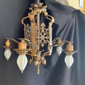 Puede incluir: Lujoso candelabro antiguo con acabado bronce. El candelabro presenta cinco lámparas con bombillas blancas en forma de lágrima. El diseño incluye intrincados adornos y elementos decorativos, suspendidos de una cadena.