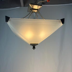 May include: A modern ceiling light fixture with a frosted glass shade in a trapezoid shape. The fixture has a silver metal frame and is suspended by chains. The corners of the shade are accented with black geometric details.