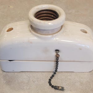 May include: A vintage white ceramic light fixture with a pull chain. The fixture has a cylindrical socket and a rectangular base with two small holes. The pull chain is a dark gray color.