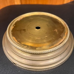 May include: A round, antique brass object with a central hole. The object has a decorative beaded edge and a slightly tarnished appearance, suggesting age and use. The base is a light gold color.
