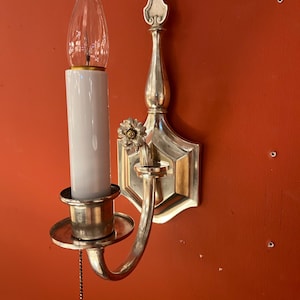 May include: A silver-toned wall sconce with a white candle-shaped light bulb. The sconce features a decorative floral accent and a pull chain. The fixture is mounted on a hexagonal backplate.