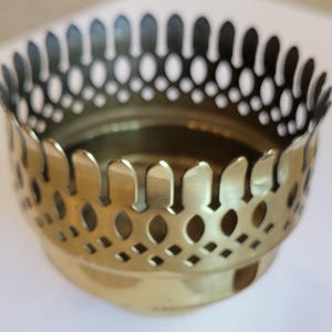 May include: A brass-colored, decorative bowl with a detailed, openwork rim. The rim features a series of oval and diamond-shaped cutouts, creating a patterned design. The bowl has a rounded base and a smooth, polished finish.