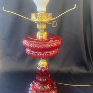 May include: A vintage red glass lamp with brass accents. The lamp features a round, patterned base and a bulbous, decorative shade. The lamp has a white light bulb and two brass arms extending from the top. The lamp is approximately 46 cm tall.