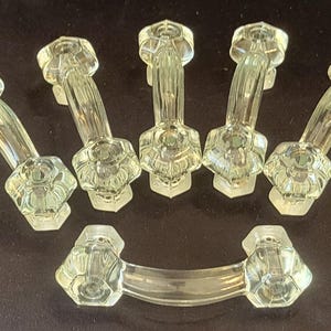 May include: A set of six clear glass drawer pulls, each featuring a curved handle and two hexagonal mounting bases with faceted edges. The handles have a smooth, transparent finish and are arranged in a group of five upright and one horizontal.