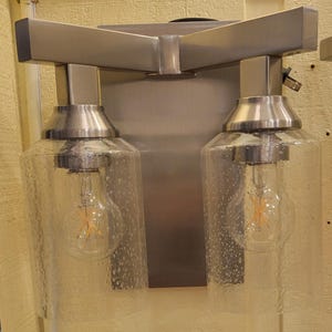 May include: A dual-light wall sconce featuring a brushed nickel finish metal frame and two cylindrical clear glass shades with a seeded texture. Each shade houses a single exposed filament light bulb, mounted on a rectangular backplate.