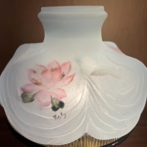 Könnte beinhalten: Ein Lampenschirm aus mattiertem Glas mit floralem Design. Der Schirm ist hellblau mit einer rosa und grünen Blume auf der Vorderseite. Der Sockel ist aus goldfarbenem Metall.
