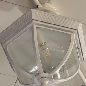 Pode incluir: Luminária externa branca com um design quadrado com painéis de vidro. A luminária tem um topo decorativo com um padrão texturizado e uma única lâmpada no interior. A luminária é montada em uma superfície branca.