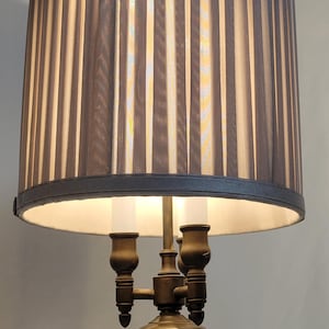 Puede incluir: Una lámpara de pie de bronce con una pantalla plisada de color gris oscuro. La pantalla tiene rayas verticales y un borde oscuro. La lámpara tiene una base decorativa y tres luces de vela falsas.