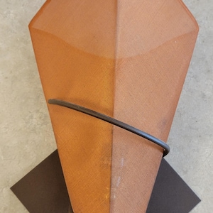May include: A geometric wall sconce with a diamond-shaped shade in shades of orange and brown. A dark metal ring wraps around the shade, and it is mounted on a dark brown square base. The sconce is a decorative lighting fixture.