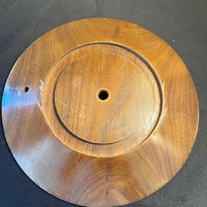 May include: A round, wooden object with a dark brown finish. The object has a raised, circular center with a hole in the middle. The outer edge is also raised, creating a tiered effect. The wood grain is visible.