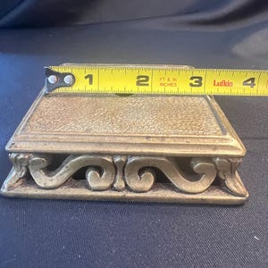 May include: A rectangular brass display stand with a textured top surface and ornate, curved scrollwork along the sides. A yellow Lufkin measuring tape is positioned across the top, indicating a width of approximately 4 inches for the base.