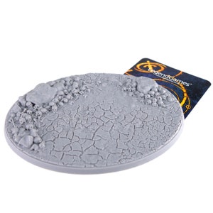 May include: A grey, oval-shaped, plastic base with a cracked, dry, desert-like texture. The base is decorated with small, skull-like shapes and rocks. The base is for use with miniature figures and games. Legend Games is printed on a card in the background.