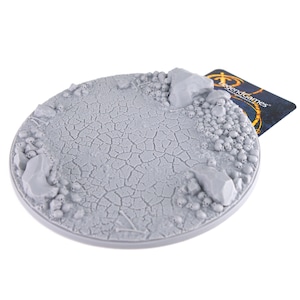 May include: A grey plastic round base with a cracked earth texture and a border of skulls and rocks. The base is designed for use with miniature figures and games.