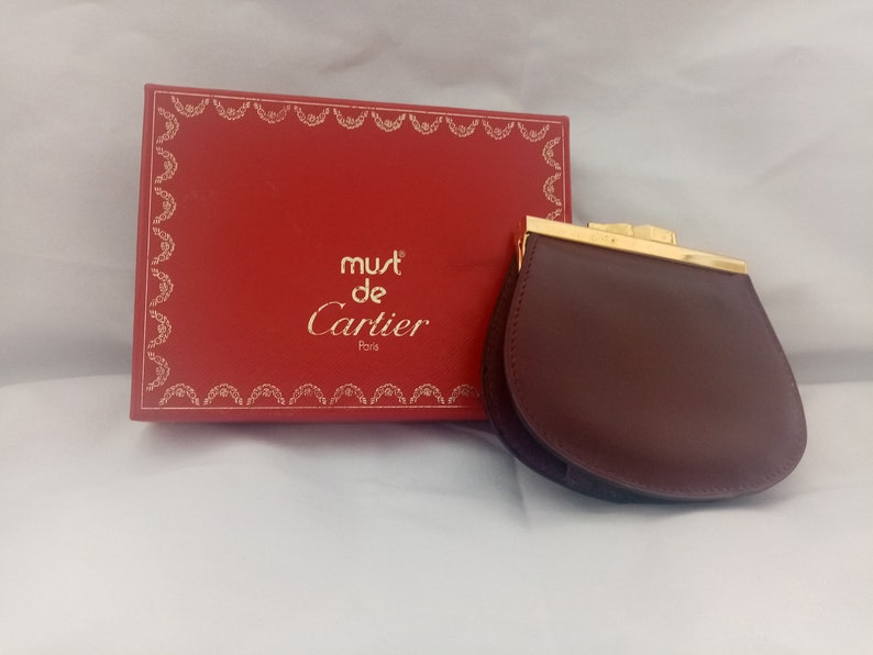 May include: A burgundy leather coin purse with a gold clasp. The purse is sitting on a white surface next to a red box with the words 'must de Cartier Paris' printed on it.