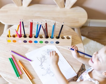 Può includere: Un bambino è seduto a un tavolo di legno con sedie a forma di coniglio, colorando con matite colorate. Il tavolo ha un porta matite incorporato con matite colorate e una fila di barattoli di vernice. Il bambino sta colorando un disegno di un pesce.