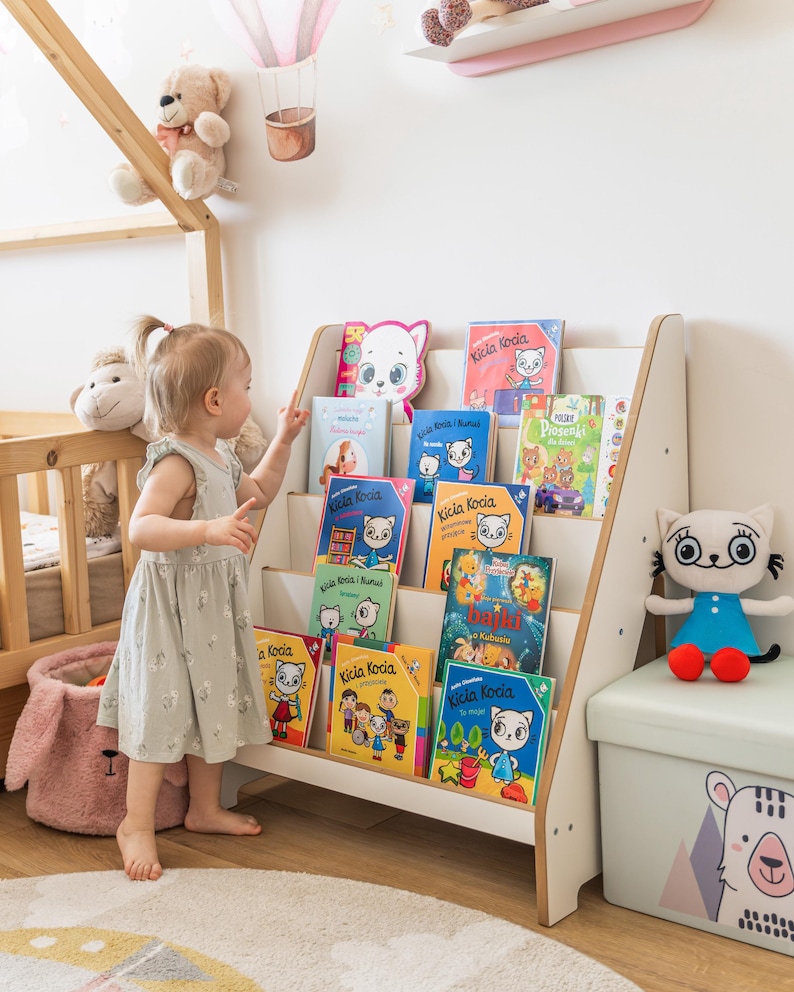 Pode incluir: Um quarto infantil com uma estante branca cheia de livros infantis coloridos. Uma crian&ccedil;a pequena com um vestido claro est&aacute; perto da estante, apontando para os livros. Um brinquedo de gato de pel&uacute;cia e uma caixa de armazenamento tamb&eacute;m s&atilde;o vis&iacute;veis.