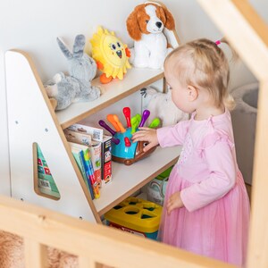Pode incluir: Uma estante de madeira para crian&ccedil;as cheia de brinquedos e livros. As prateleiras cont&ecirc;m um coelho de pel&uacute;cia cinza, um sol amarelo, um cachorro de pel&uacute;cia e um classificador de formas. Uma crian&ccedil;a com um vestido rosa alcan&ccedil;a um brinquedo.