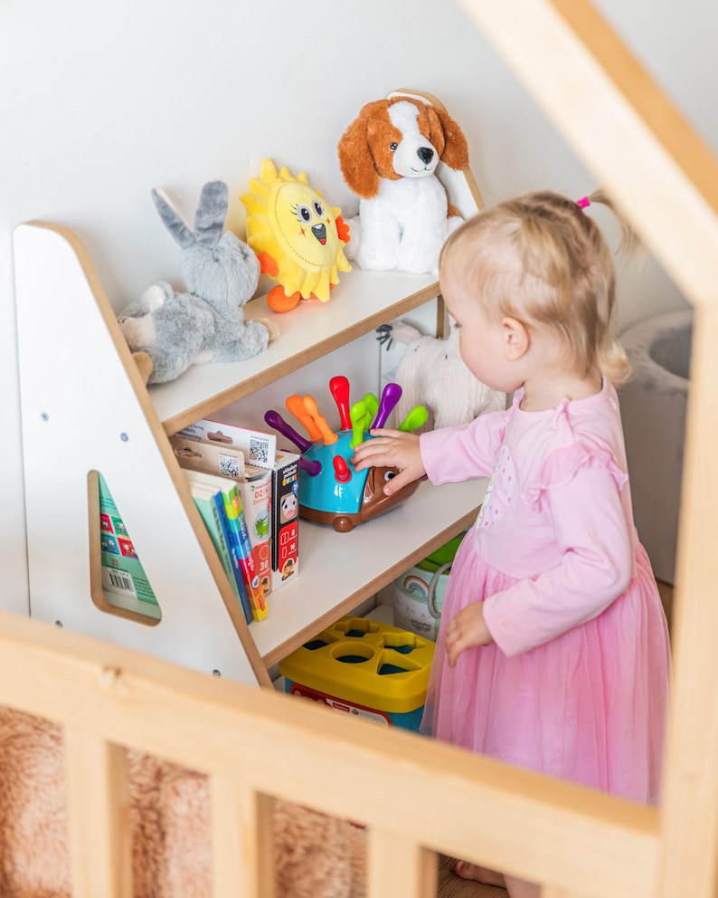 Pode incluir: Uma estante de madeira para crian&ccedil;as cheia de brinquedos e livros. As prateleiras cont&ecirc;m um coelho de pel&uacute;cia cinza, um sol amarelo, um cachorro de pel&uacute;cia e um classificador de formas. Uma crian&ccedil;a com um vestido rosa alcan&ccedil;a um brinquedo.
