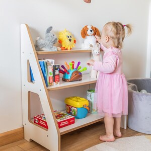 Pode incluir: Uma estante infantil branca e de madeira com tr&ecirc;s prateleiras, cheia de brinquedos e livros. Um coelho de pel&uacute;cia cinza, um sol amarelo e um c&atilde;o de pel&uacute;cia s&atilde;o vis&iacute;veis. Uma crian&ccedil;a pequena com um vestido rosa alcan&ccedil;a um brinquedo.