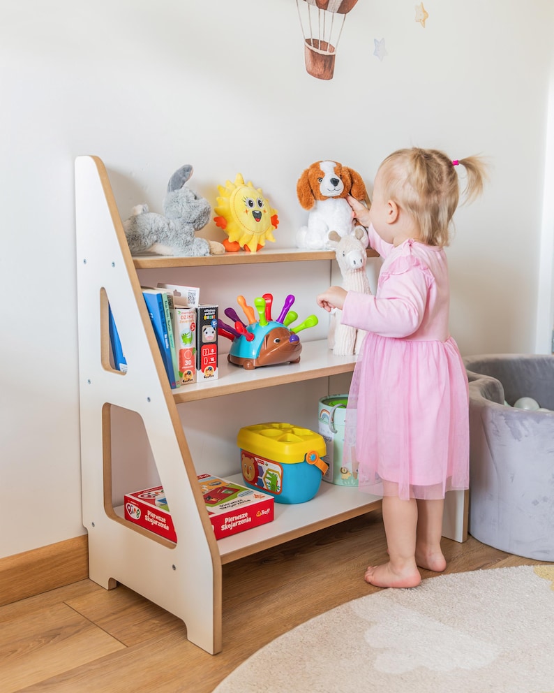 Pode incluir: Uma estante infantil branca e de madeira com tr&ecirc;s prateleiras, cheia de brinquedos e livros. Um coelho de pel&uacute;cia cinza, um sol amarelo e um c&atilde;o de pel&uacute;cia s&atilde;o vis&iacute;veis. Uma crian&ccedil;a pequena com um vestido rosa alcan&ccedil;a um brinquedo.