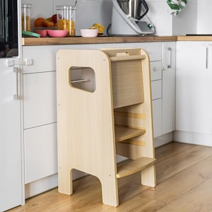 May include: A light-coloured wooden learning tower with a tall frame, a single step, and a platform. The structure features side cutouts for handles, a horizontal top bar, and a smooth finish, designed as a standing platform for elevated kitchen access.