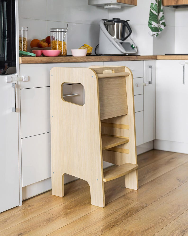 May include: A light-coloured wooden learning tower with a tall frame, a single step, and a platform. The structure features side cutouts for handles, a horizontal top bar, and a smooth finish, designed as a standing platform for elevated kitchen access.